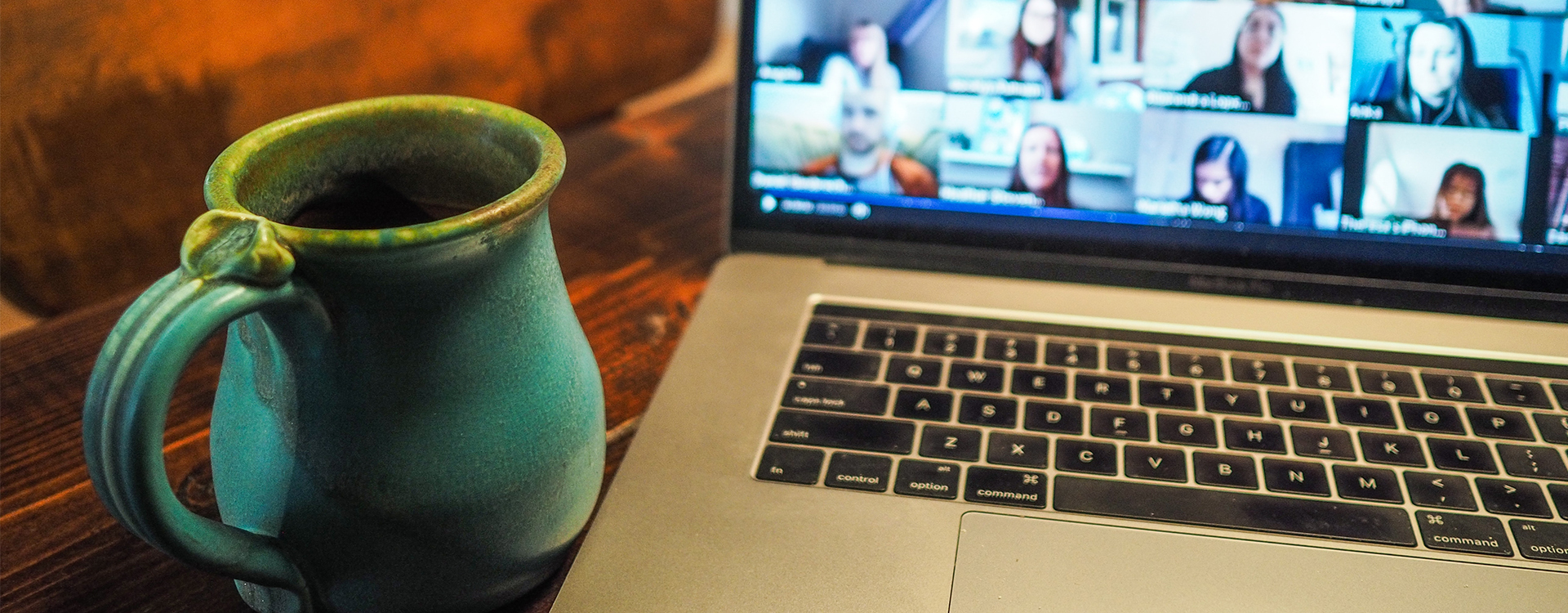 Ein Tisch mit einem Laptop drauf, dessen Bildschirm die Teilnehmer eines digitalen Meetings zeigt. Links neben dem Computer steht eine grüne Tasse.