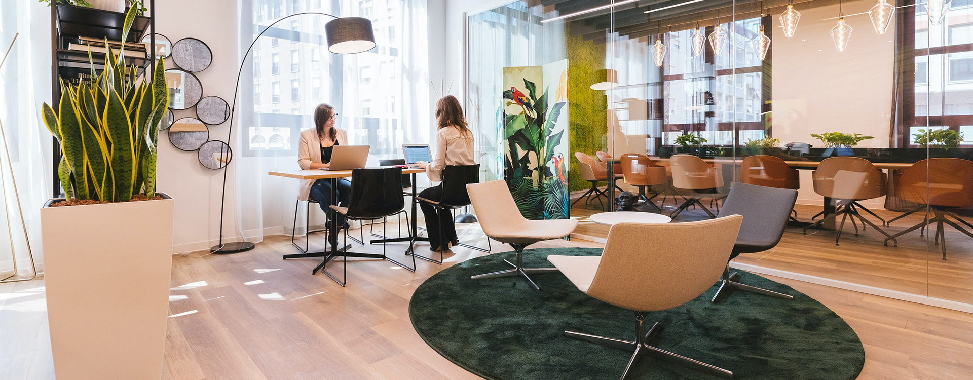 To kvinder sidder og arbejder på deres bærbare computere ved et bord. I forgrunden ses tre loungestole omkring et sofabord på et grønt tæppe. Et møbleret mødelokale ses i baggrunden.